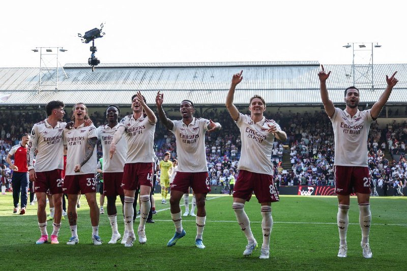 Seven soccer players in white jerseys and maroon shorts celebrate with raised arms on a green pitch, with a stadium crowd and a television camera visible in the background.