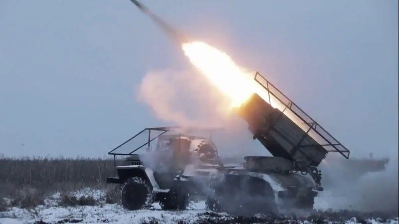 A military truck-mounted rocket launcher fires a rocket, producing a bright flame and smoke plume in a snowy field.