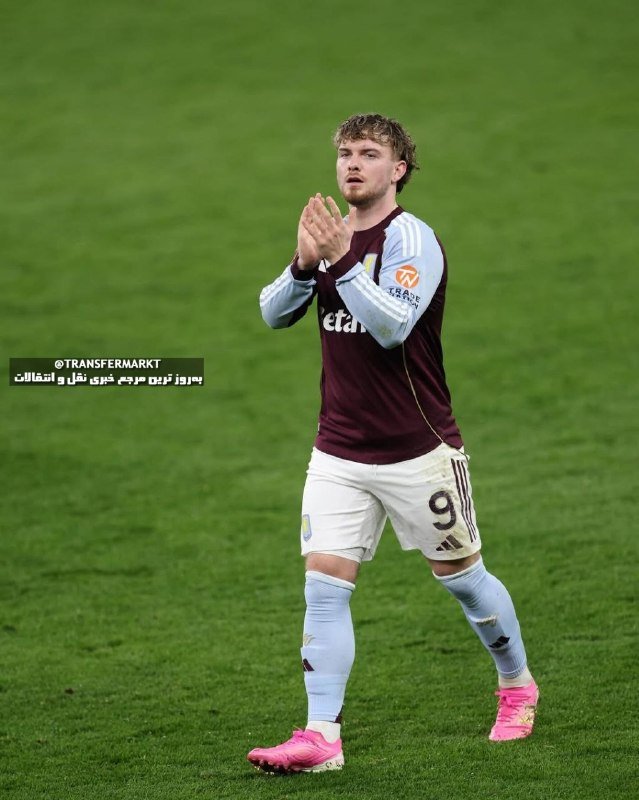 A footballer in a claret and blue jersey with the number 9 claps his hands while standing on a grass pitch, wearing pink boots.