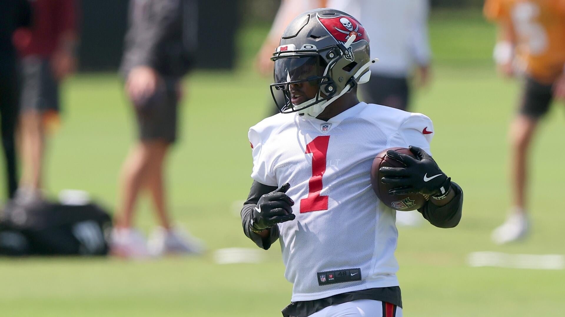 A football player wearing a white jersey with the number 1 and a Tampa Bay Buccaneers helmet runs with the ball during a daytime practice on a grass field.