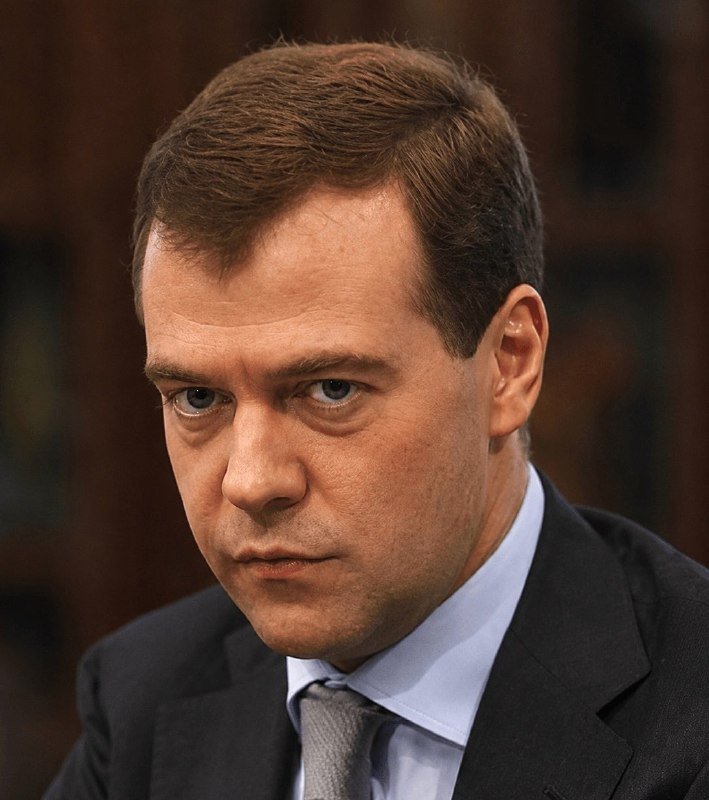 A man in a dark suit, light blue shirt, and gray tie looks forward with a serious expression against a blurred dark background.