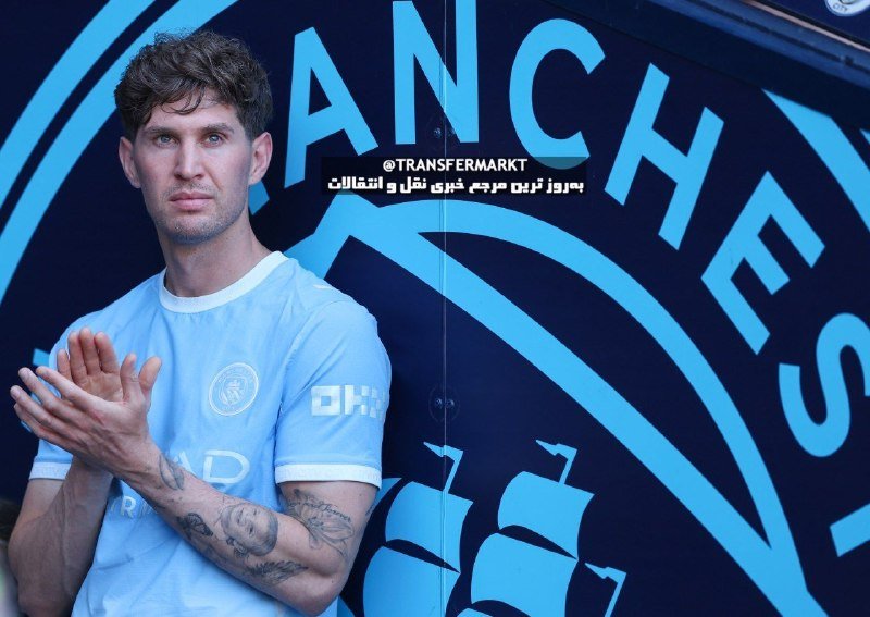 A man in a light blue Manchester City jersey stands clapping in front of a large Manchester City backdrop featuring the club's crest and anchor design.