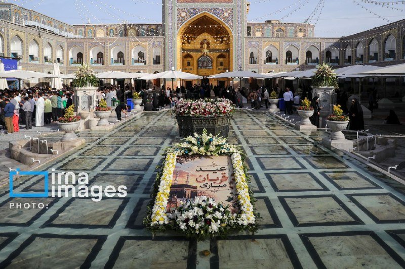 Crowds gather in a tiled courtyard of an ornate shrine with a gilded archway, where a floral display featuring Persian script surrounds a framed picture on the ground.