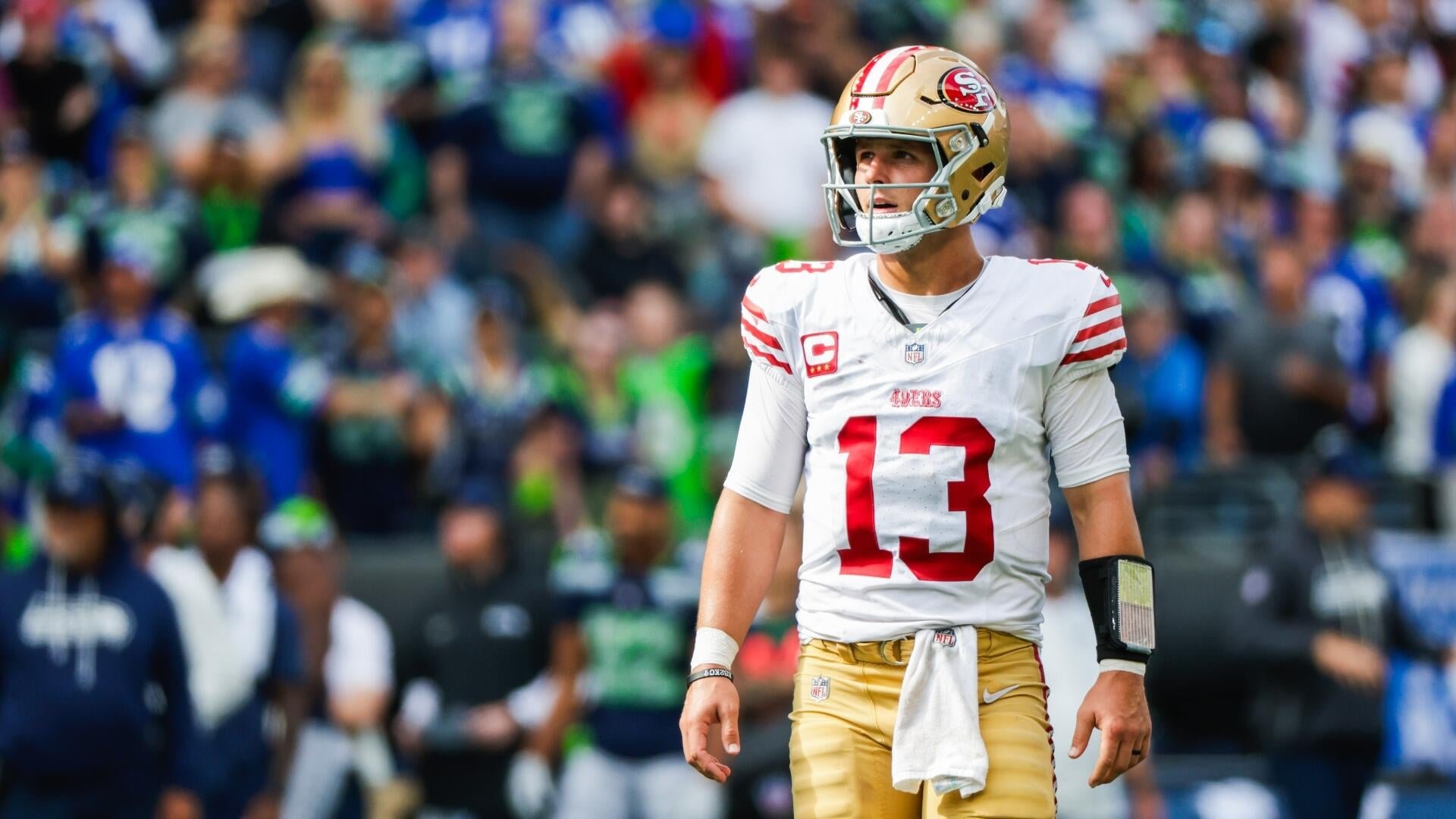 San Francisco 49ers quarterback wearing jersey number 13 walks off the field during an NFL game, with a blurred crowd visible in the stadium background.