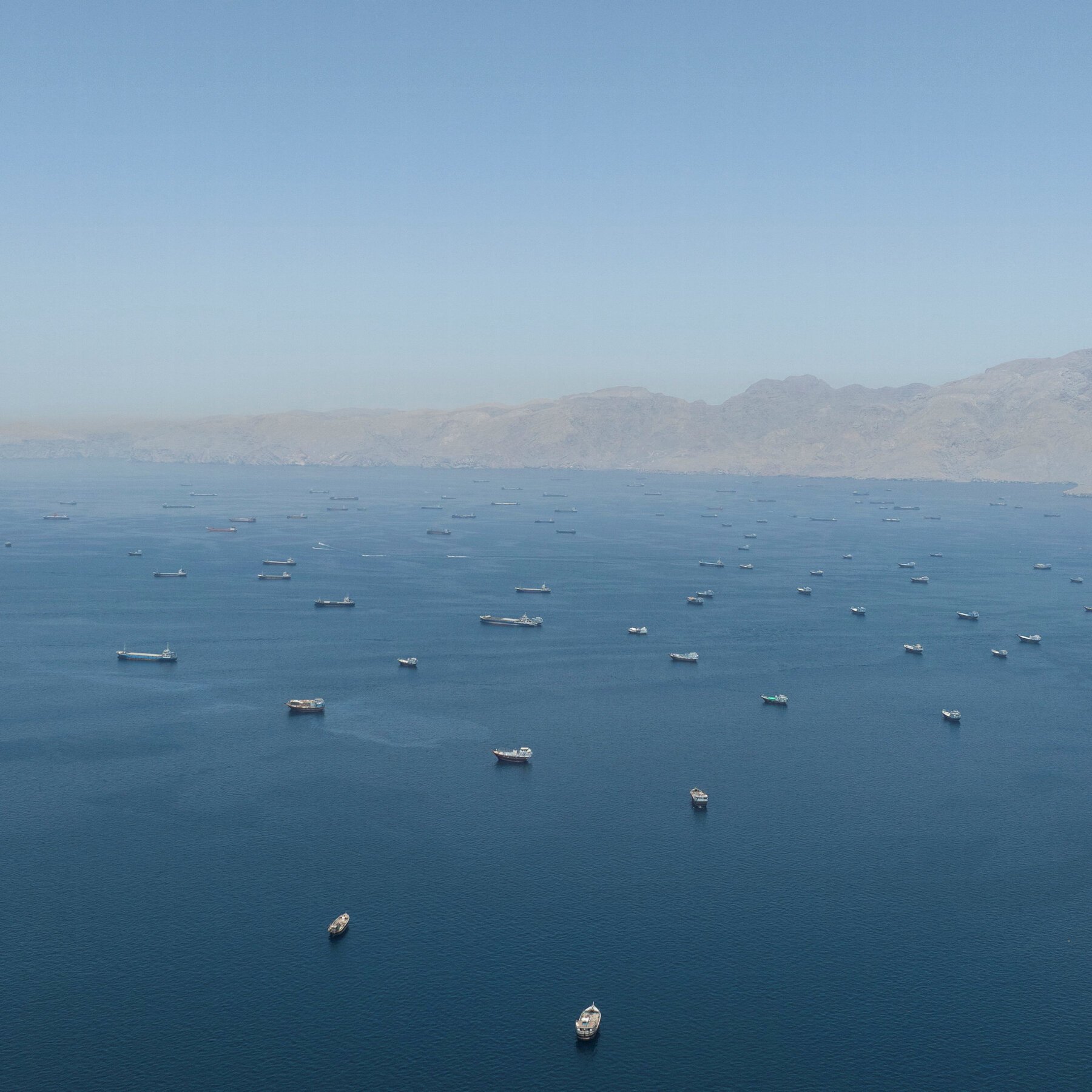 Dozens of cargo ships and vessels are anchored in a calm blue sea, with a hazy mountain range visible in the background under a clear sky.