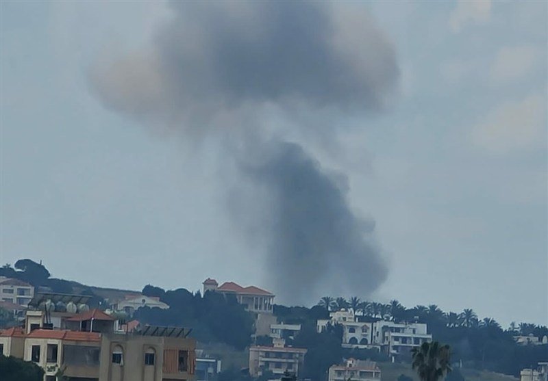 A large plume of dark gray smoke rises into a pale sky above a coastal town with low-rise buildings, red-roofed houses, and scattered palm trees.