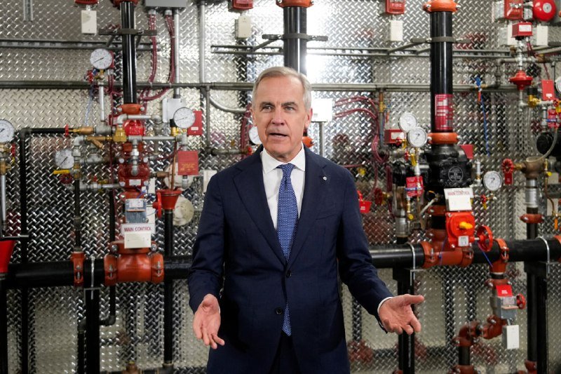 A man in a dark suit and blue tie stands with arms outstretched in front of an industrial system of red pipes, valves, and pressure gauges.