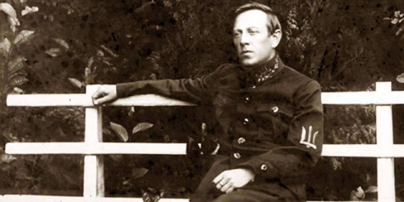 A sepia-toned photograph shows a man in a dark military-style jacket leaning against a white wooden fence, with foliage visible in the background.