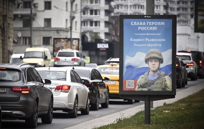 A street-side billboard displaying a Russian-language poster with a soldier's portrait stands beside a line of cars in traffic on a city street.