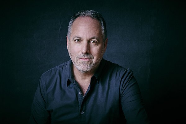 A gray-haired man in a dark button-up shirt poses for a portrait against a black background.