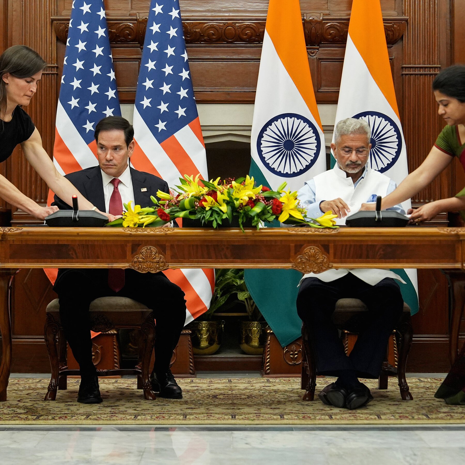 Two men in suits sit at an ornate wooden table signing documents, flanked by U.S. and Indian flags, with floral arrangements and two assistants in the background.