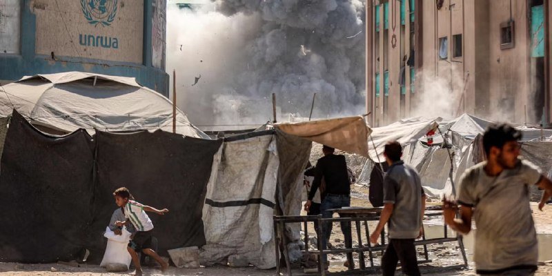 A plume of dark smoke rises behind a UNRWA-branded building and makeshift tents, as people, including a child carrying a bag, walk through the dusty area.