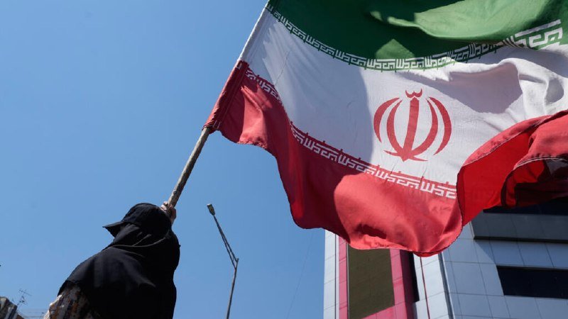 A person wearing a black head covering waves a large Iranian flag against a clear blue sky.