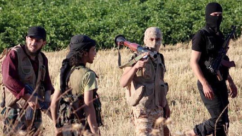 Four masked and armed individuals in tactical vests walk through a dry grassy field, one carrying a shoulder-fired weapon.