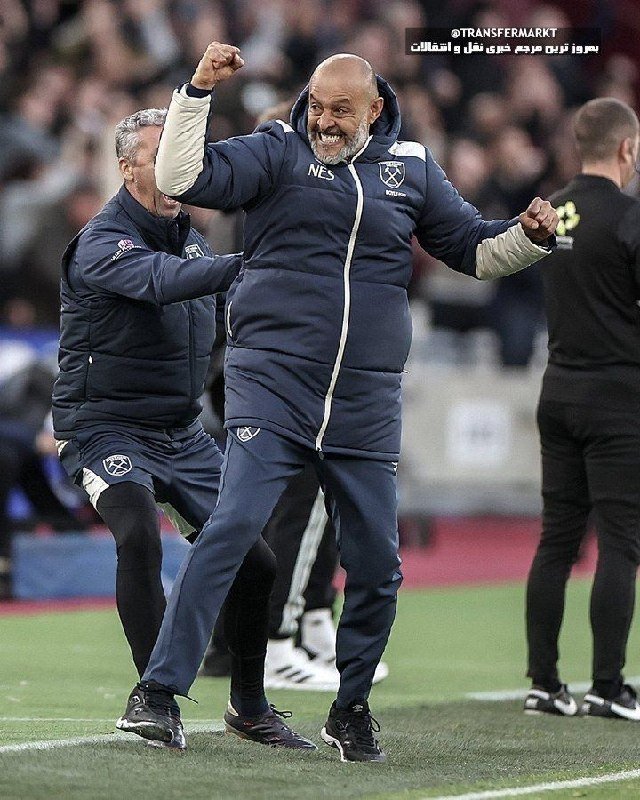 Two men in dark West Ham tracksuits celebrate with raised fists on a football pitch sideline, with a blurred crowd visible in the background.