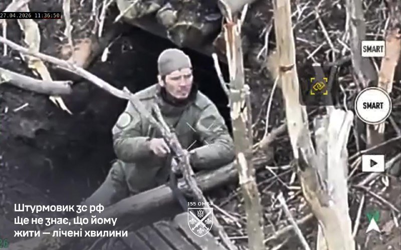 A soldier in camouflage uniform and knit cap sits holding a rifle in a trench filled with broken branches and debris.