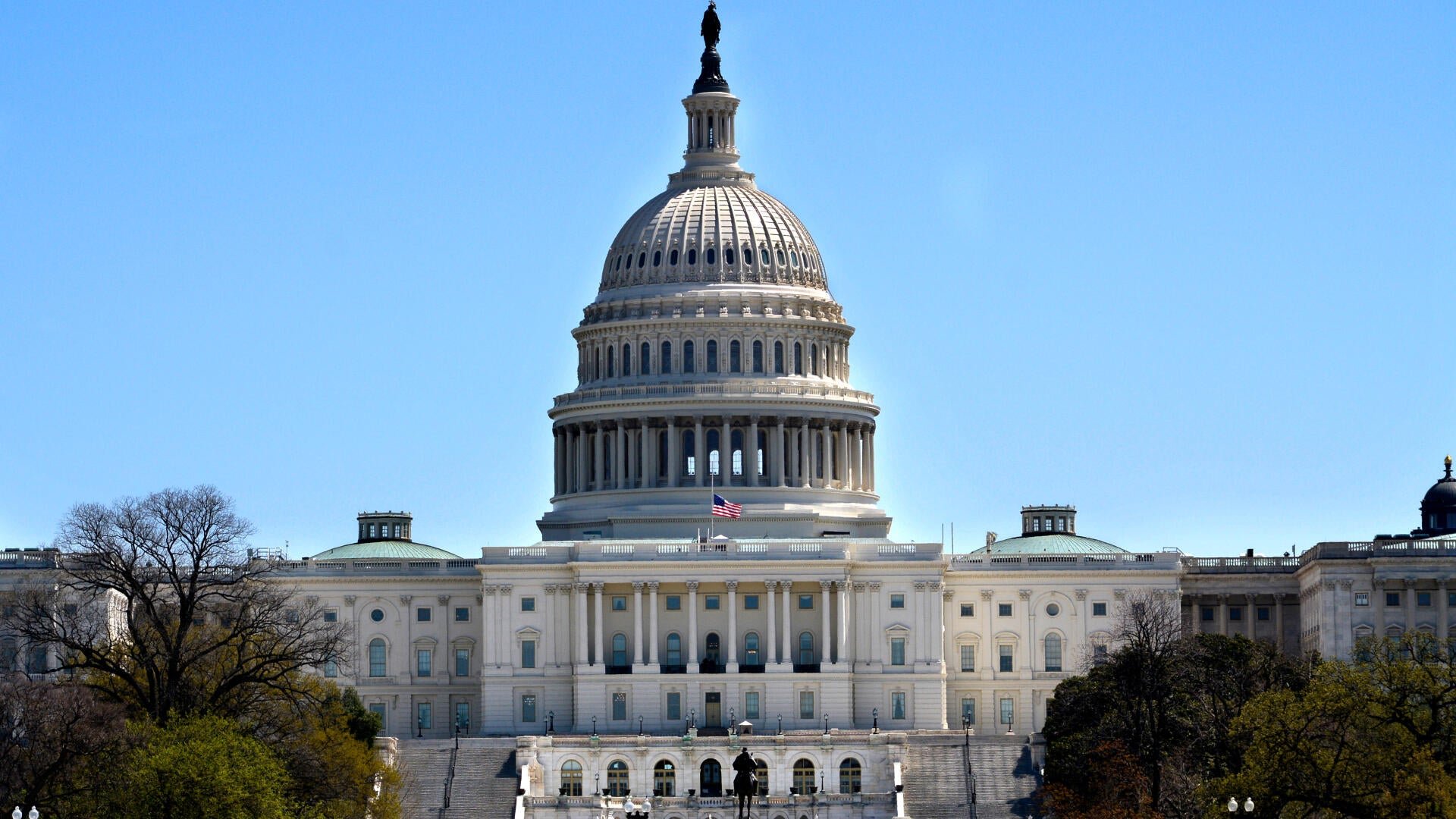 The U.S. Capitol building's white domed structure stands against a clear blue sky, with an American flag flying below the dome and trees framing the foreground.