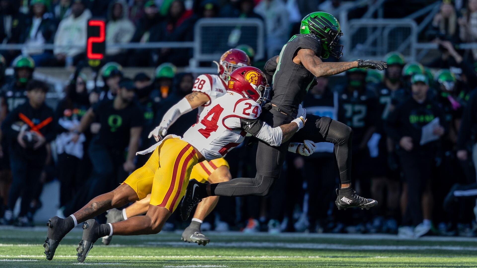 A football player in a black uniform and green helmet runs with the ball while being tackled by two opposing players in white and cardinal uniforms, with a crowd in the background.