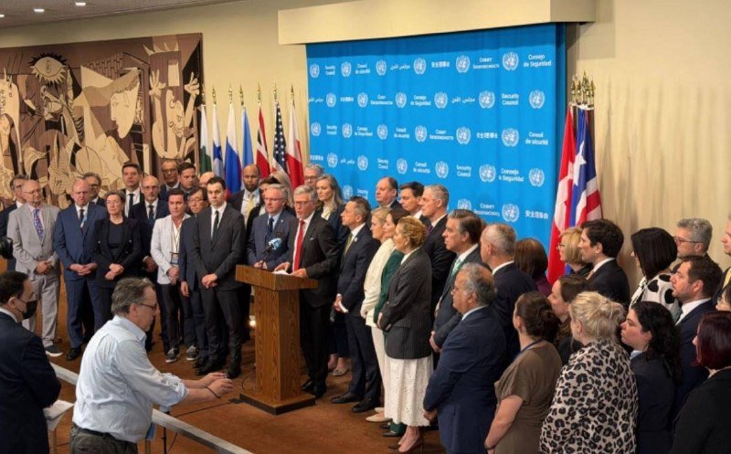 A man speaks at a wooden podium before a backdrop displaying United Nations and Security Council logos, flanked by flags, as a crowd of formally dressed people stands listening.