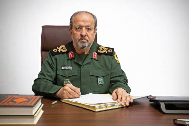 A man in a green military uniform with insignia sits at a desk, holding a pen over an open notebook beside stacked books and a laptop.