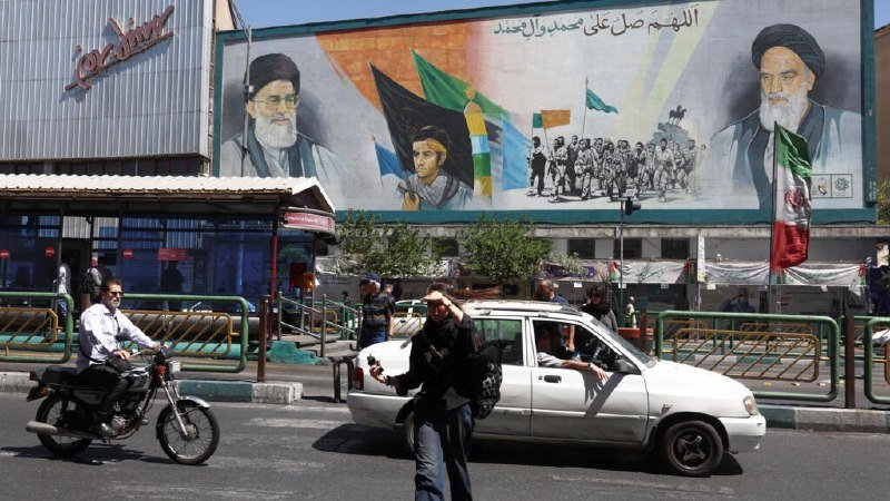 A large billboard featuring two bearded religious figures, flags, and a youth's portrait overlooks a street where pedestrians, a motorcyclist, and a white car pass by.