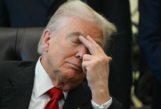 A man with blonde hair wearing a dark suit and red tie sits with his eyes closed, pressing his fingers to his forehead in an apparent gesture of fatigue or frustration.