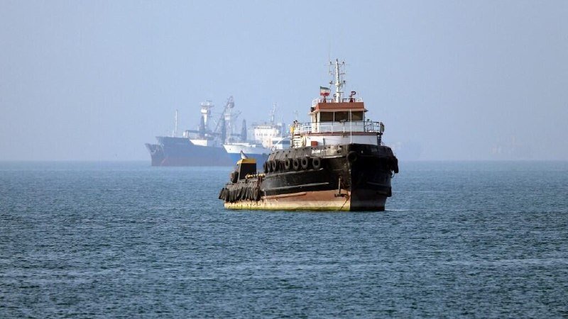 A black and white tugboat navigates calm, hazy waters in the foreground, with a larger cargo ship and industrial cranes visible in the distant background.