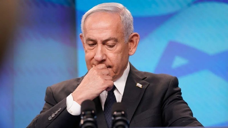 A gray-haired man in a dark suit and tie rests his fist against his mouth, standing behind two microphones against a blue backdrop featuring a partial star design.