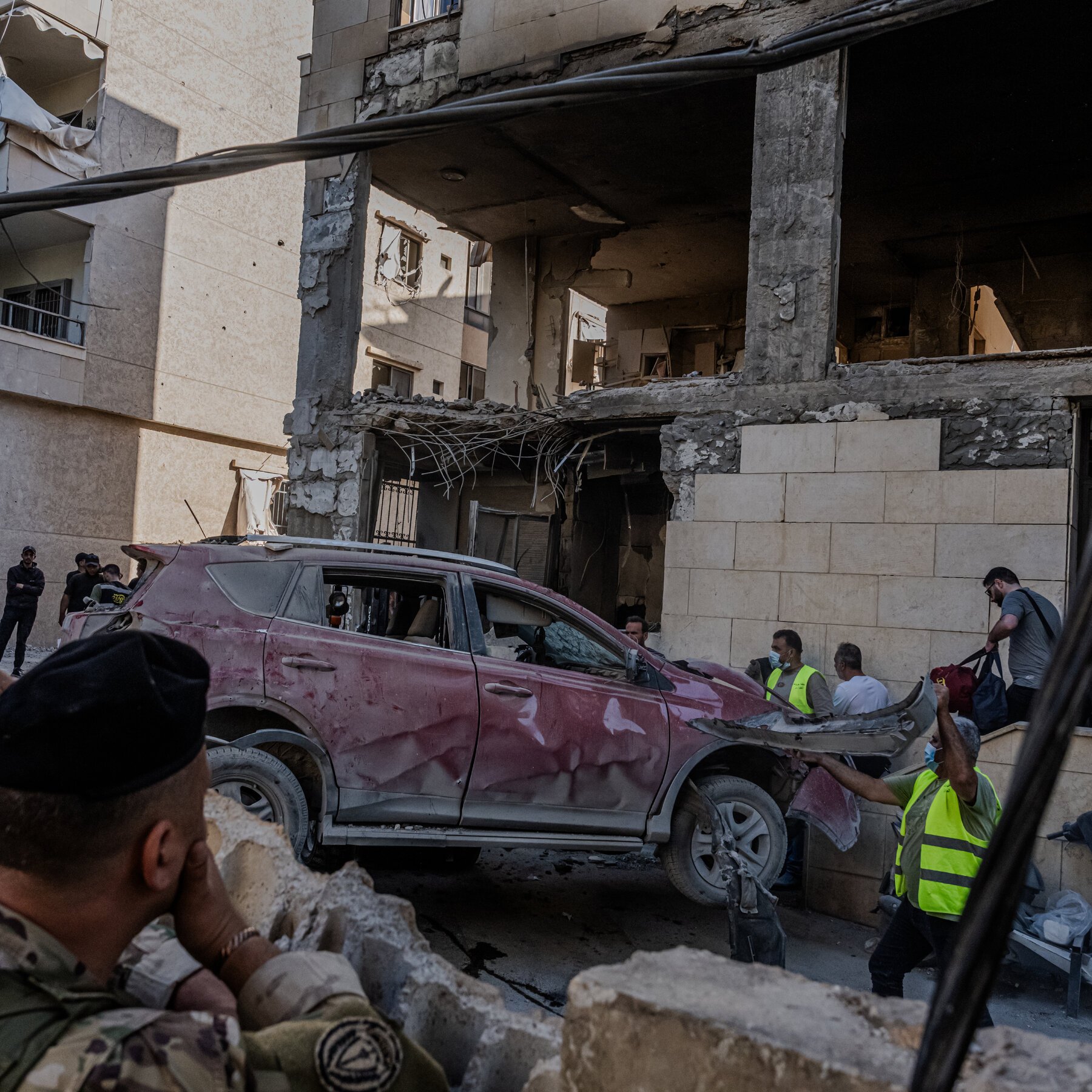 Workers in high-visibility vests inspect a damaged, dust-covered car in front of a heavily destroyed multi-story building with exposed rebar and concrete rubble.