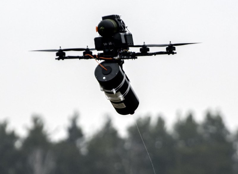 A multi-rotor drone equipped with a camera and a suspended cylindrical payload hovers mid-air above a blurred treeline.