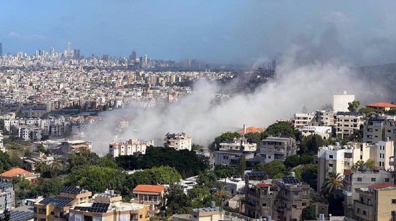 Thick gray smoke rises from a densely populated urban area, billowing over low-rise buildings with red-tiled and flat rooftops against a hazy sky.