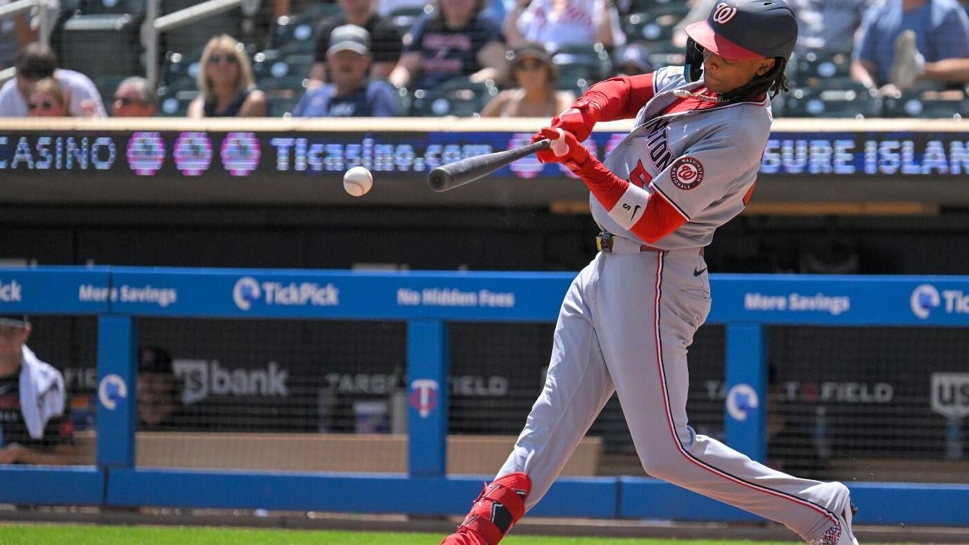 A baseball player in a gray and red uniform swings a bat at an incoming ball, with spectators and advertisements visible in the background.
