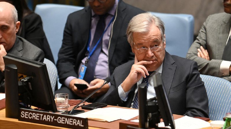 A gray-haired man in a suit sits at a desk labeled "SECRETARY-GENERAL," resting his chin on his hand, with other individuals seated behind him in a formal meeting room.