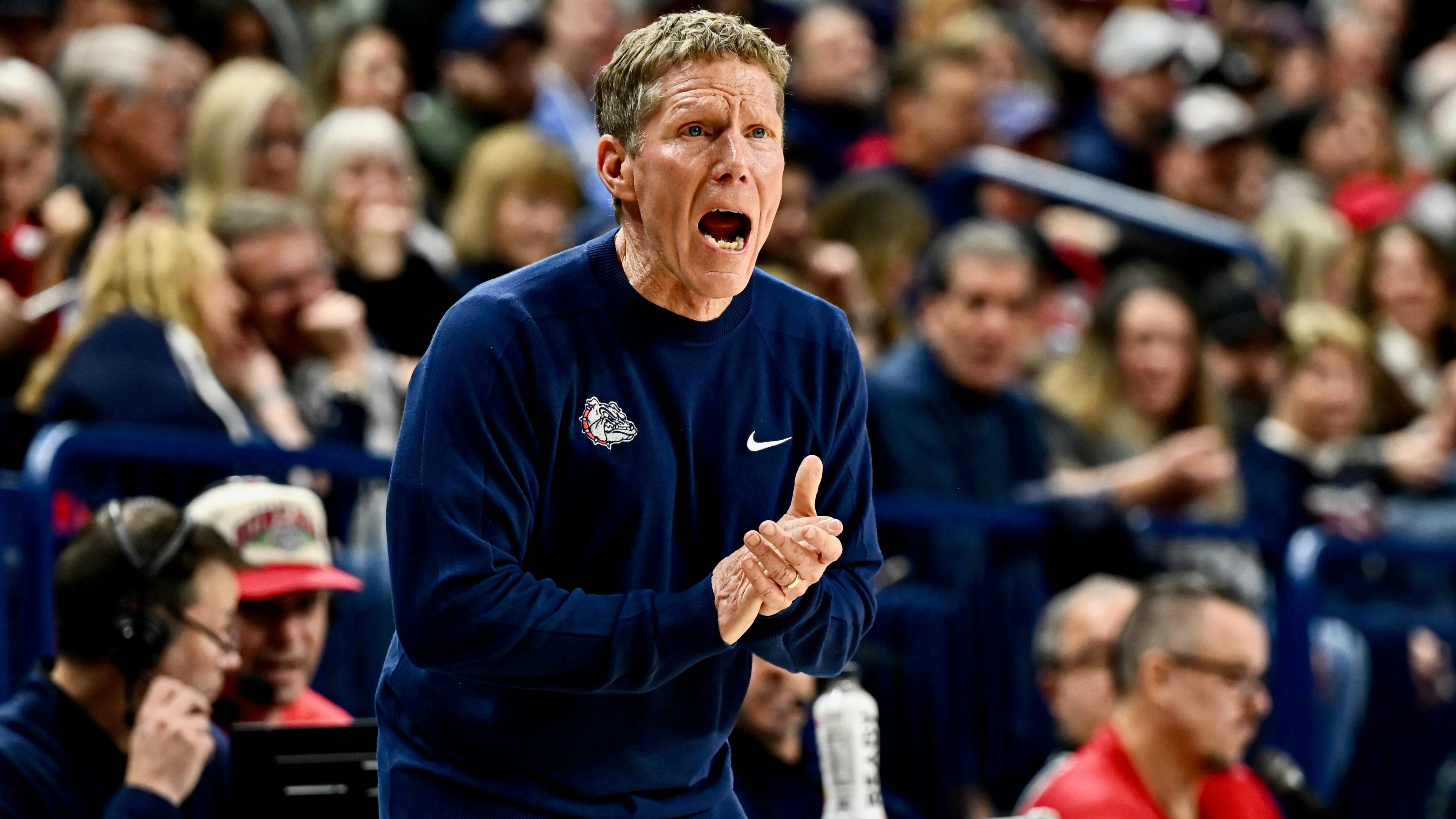 A man in a navy blue Bulldogs sweatshirt claps and shouts from the sideline during a basketball game before a crowd of spectators.
