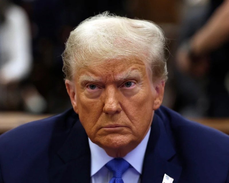 An older man with blonde hair, wearing a dark suit, white shirt, and blue tie, sits with a serious expression in a formal indoor setting.