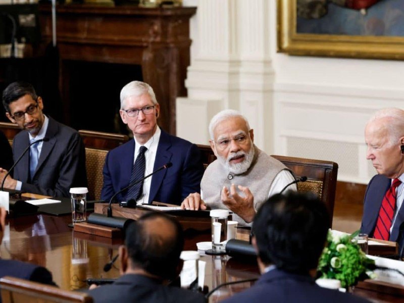 Four men in business attire sit at a polished conference table with microphones, one gesturing with his hand as if speaking, in a formal room with a fireplace and framed painting.