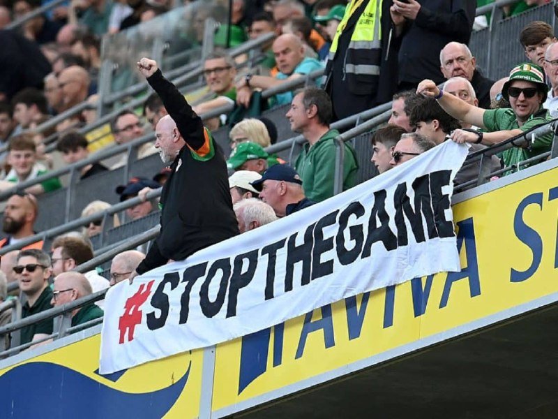 A crowd of sports fans in green attire cheer in stadium stands, with one man raising his fist above a white banner reading "#STOPTHEGAME."