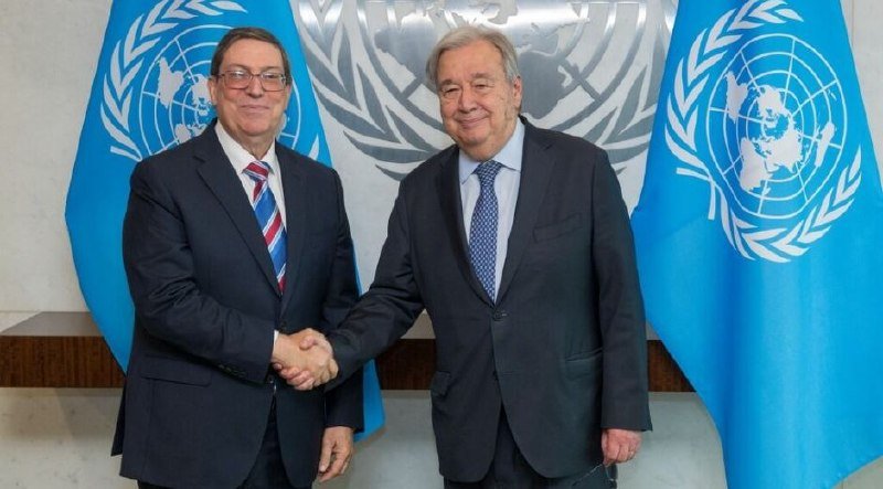 Two men in dark suits shake hands and smile, standing before a UN emblem and two light-blue UN flags.