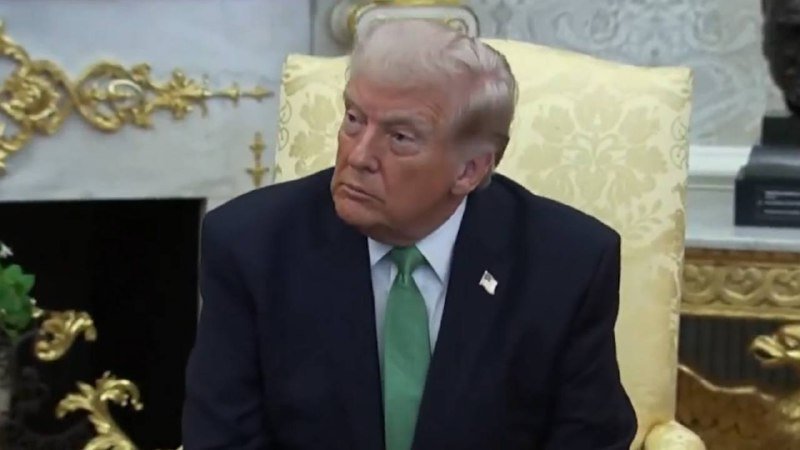 A man in a dark suit and green tie sits in an ornate yellow chair in a gilded, formal room.
