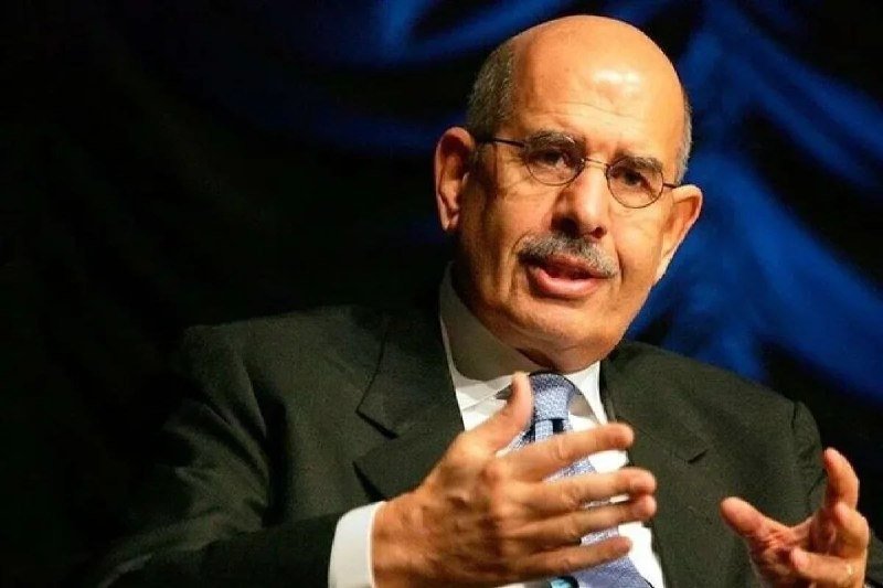 A bald, mustachioed man in a dark suit and blue tie gestures with both hands while speaking against a dark blue backdrop.