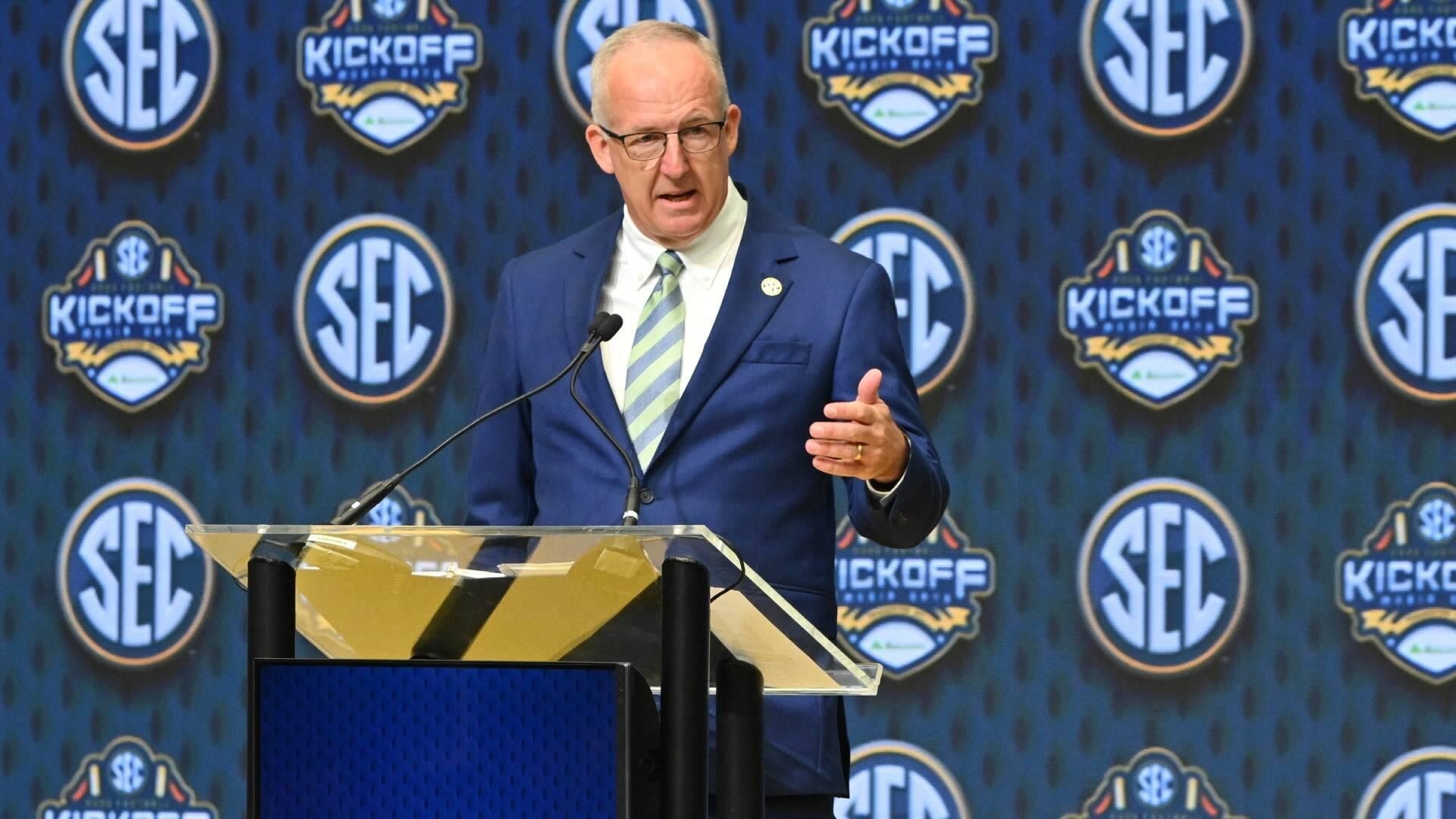 An older man in a blue suit and green-striped tie speaks at a clear podium, gesturing with one hand, against a backdrop displaying SEC and SEC Kickoff logos.