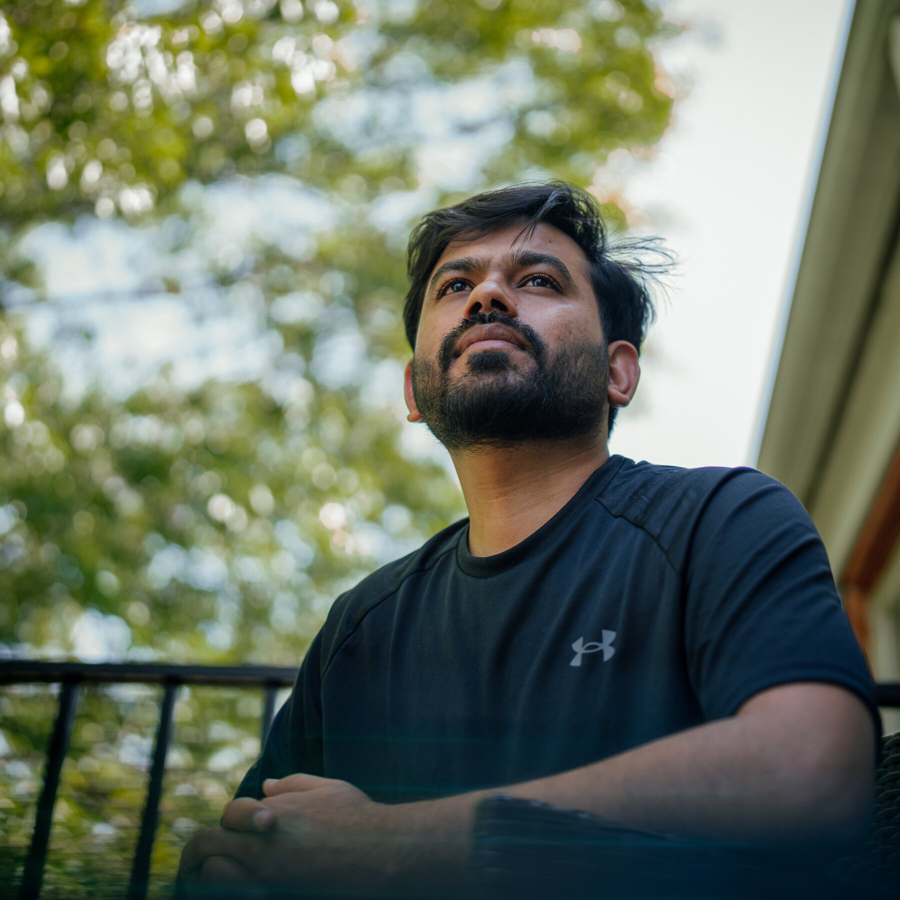 A bearded man wearing a black Under Armour t-shirt looks upward while seated on a balcony with a metal railing, green trees visible in the background.
