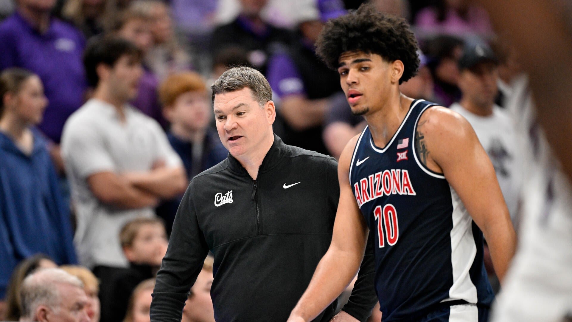A basketball coach in a black "Cats" quarter-zip walks alongside a player wearing an Arizona Wildcats navy jersey with the number 10, with a crowd visible in the background.