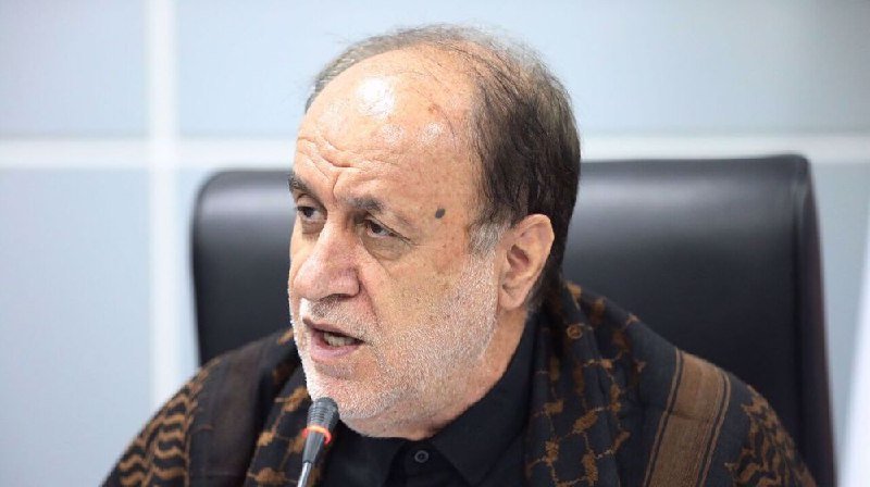 An older man with gray hair and a patterned shawl speaks into a microphone while seated in a black office chair.