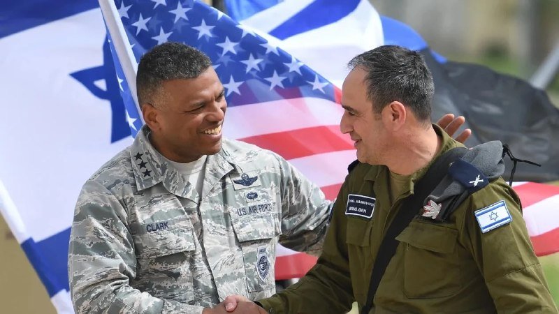 Two military officers—one in a U.S. Air Force uniform labeled "Clark," the other in an Israeli uniform—shake hands and smile in front of American and Israeli flags.