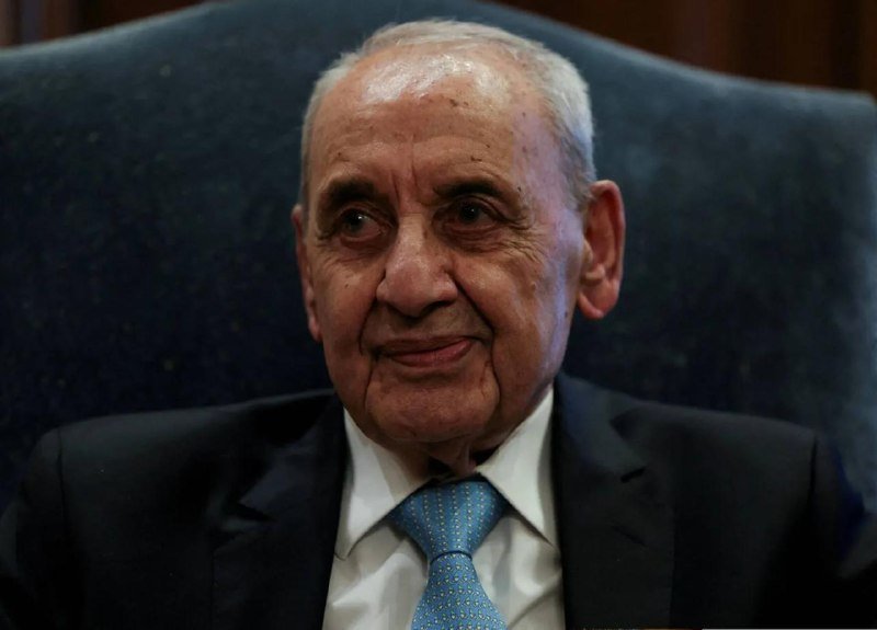 An elderly man in a dark suit and light blue tie sits in a high-backed blue chair, looking off to the side with a slight smile.