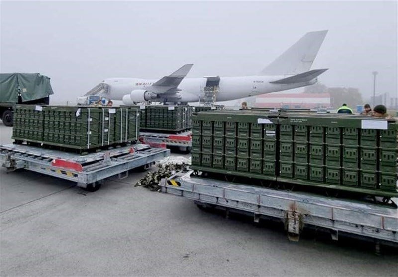 Green military crates stacked on flatbed dollies sit on a foggy tarmac in front of a parked white cargo aircraft with a loading ramp extended.