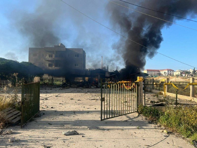 Thick black smoke rises from a burning debris-filled lot enclosed by a metal gate, with residential buildings visible in the background under a clear sky.