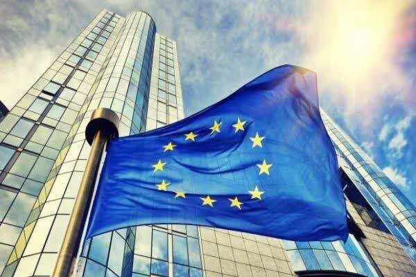 European Union flag waves in front of a modern glass skyscraper against a sunny blue sky.