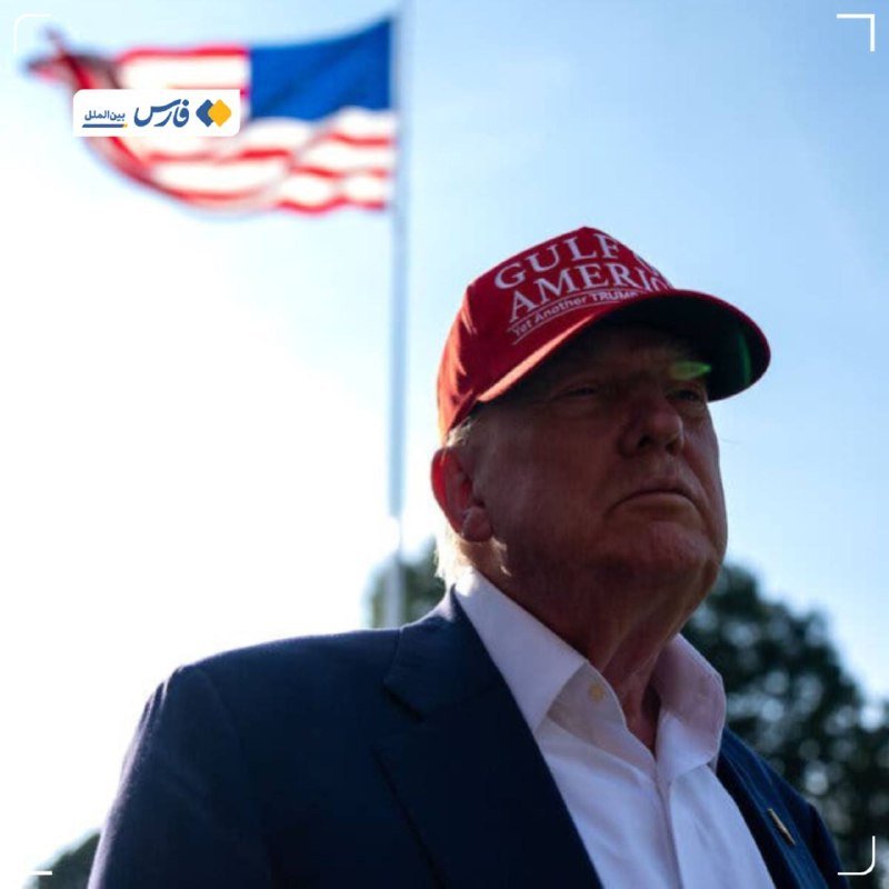 A man in a navy blazer and white shirt wears a red "GULF OF AMERICA, 1st Annual Trump" cap, with an American flag waving in the background.